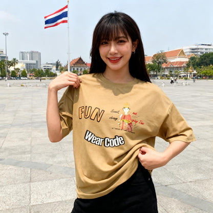 Woman wearing a beige t-shirt with 'FUN Wear Code' text and a playful Thai woman graphic, standing outdoors with a Thai flag and buildings in the background.